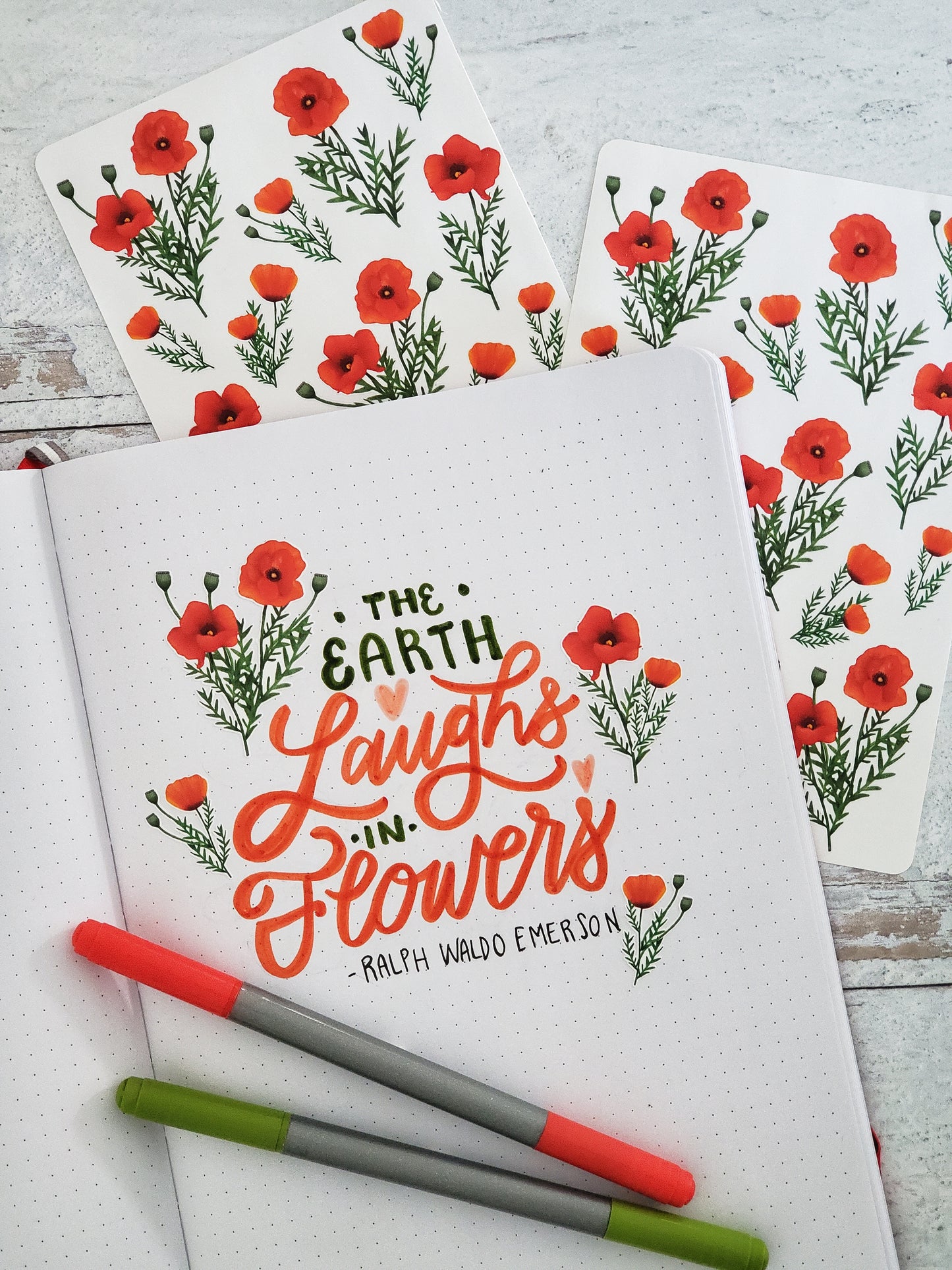 A few of the poppy stickers decorating a hand lettered quote written in a journal. The quote reads "the Earth Laughs in Flowers - Ralph Waldo Emerson"