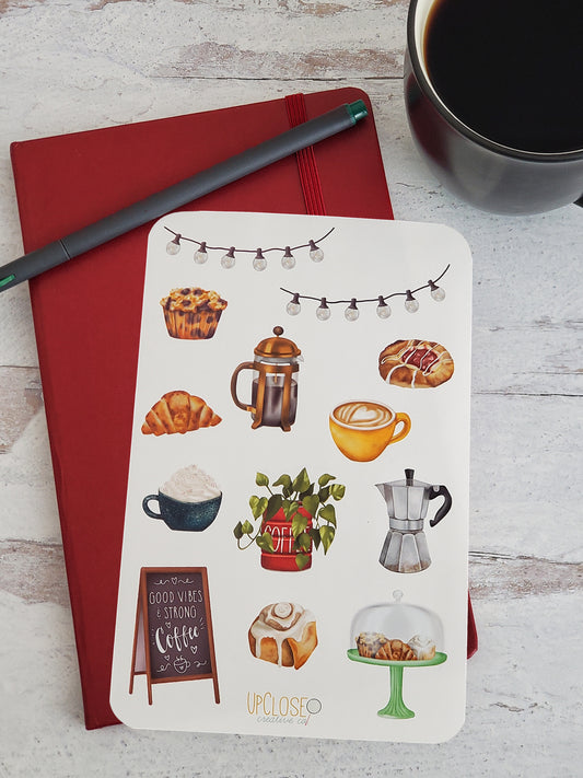 A sticker sheet featuring an assortment of illustrated items found in a coffee shop. The sticker sheet is sitting on top of a red journal next to a cup of coffee. 