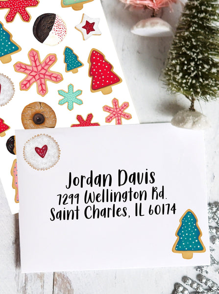 a blue frosted Christmas tree cookie and a heart shaped jam cookie decorate a white envelope surrounded by Christmas decoration